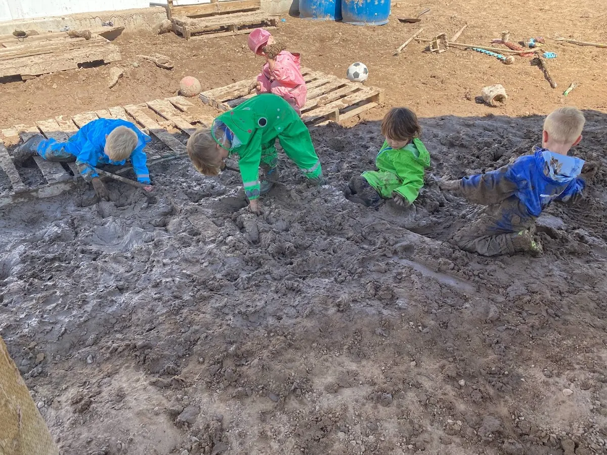 Children in colorful rain suits crawling, digging, and sitting in a wide mud pit
