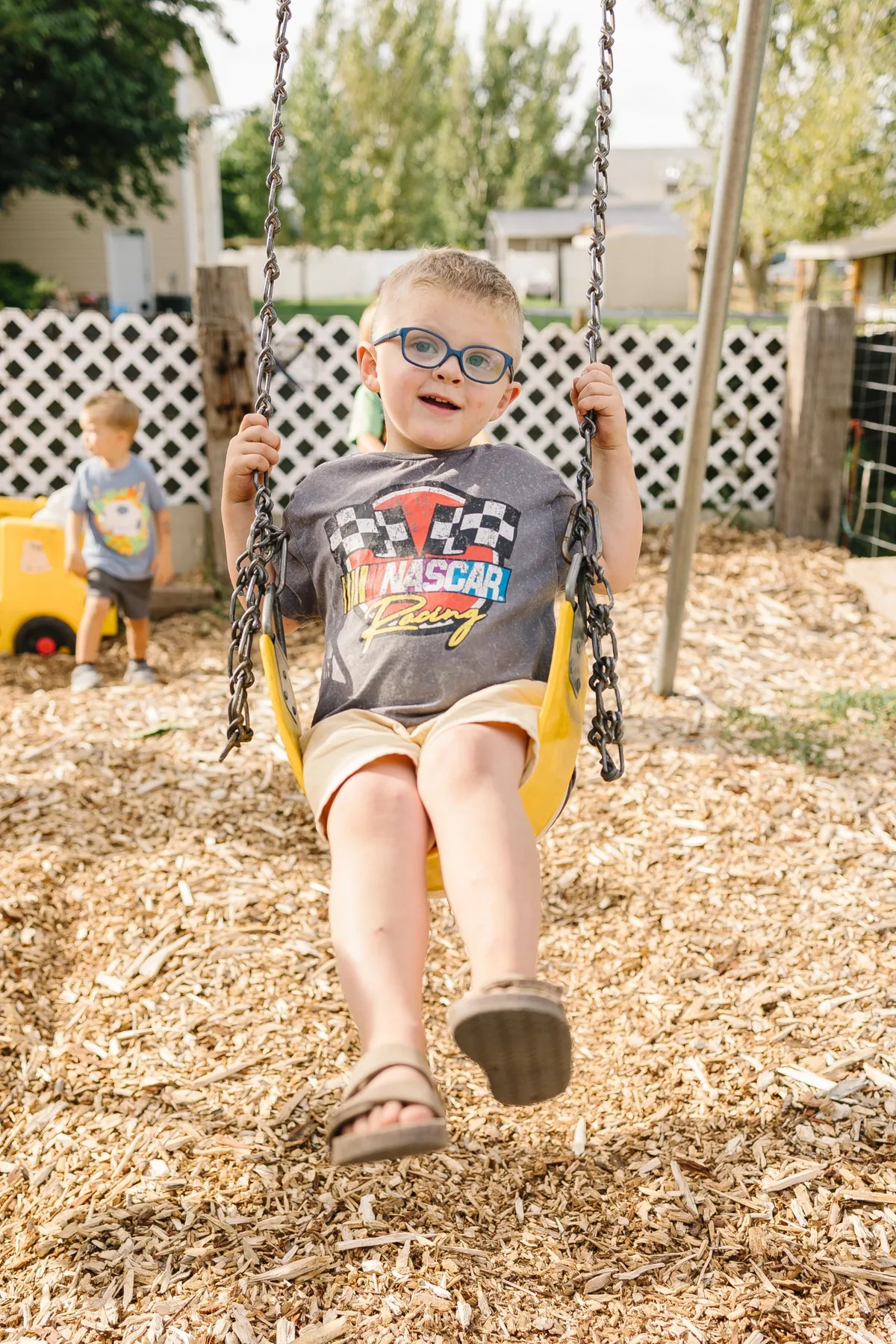 A smiling child in glasses swinging on a yellow bucket swing over the wood-chip play yard