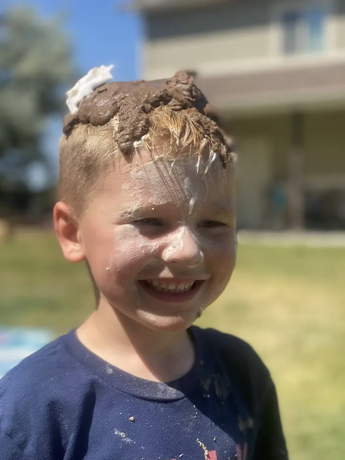 A preschooler laughing with a heap of mud piled on top of their head during outdoor sensory play