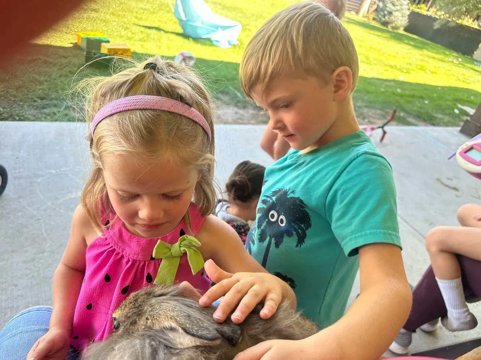 Two children gently petting a small furry animal on a shaded patio