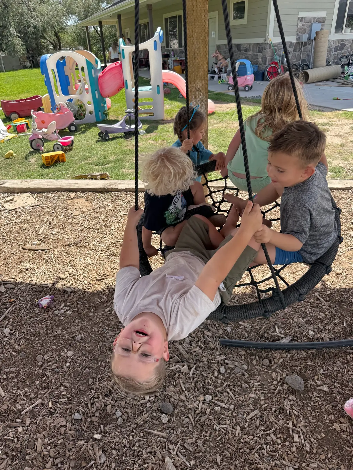 A child hanging upside down from a rope nest swing with friends aboard, in front of a grassy yard full of ride-on toys