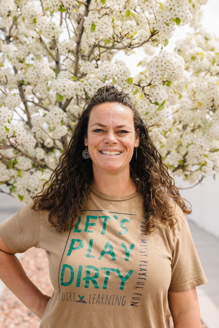 Christi Warr, founder of Dirt Learning, smiling in a “Let's Play Dirty” t-shirt beneath a blossoming tree