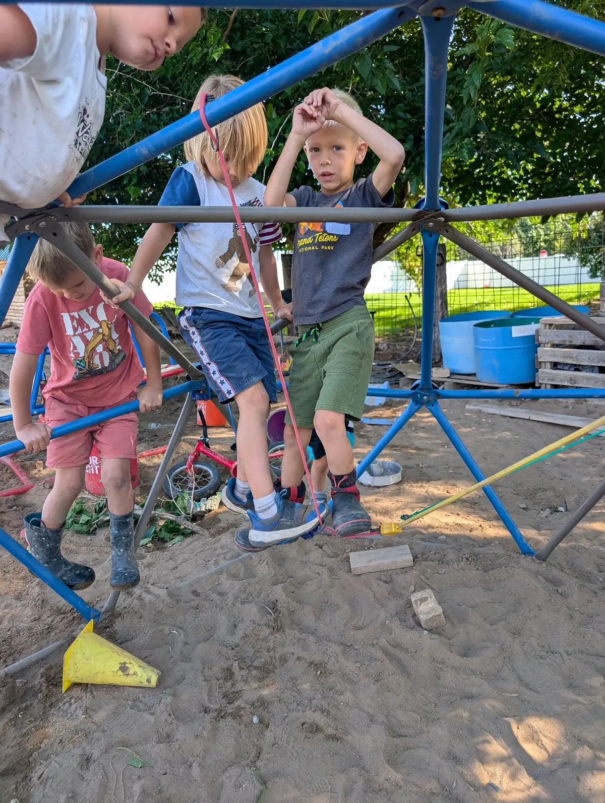 Four children climbing and balancing together on a blue geodesic climbing dome over sandy ground