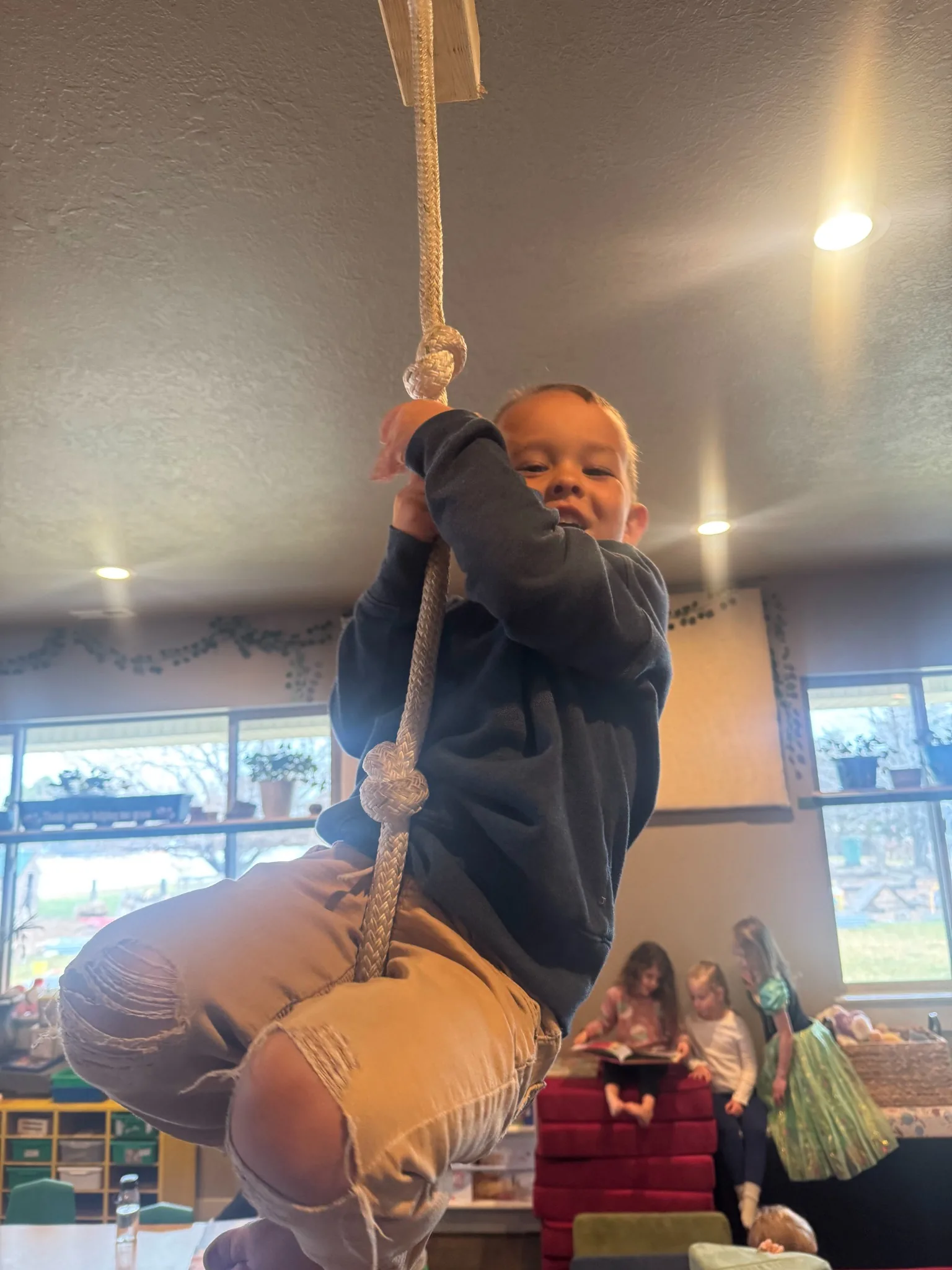 A child climbing a knotted rope hung from the indoor classroom ceiling while friends read together in the background