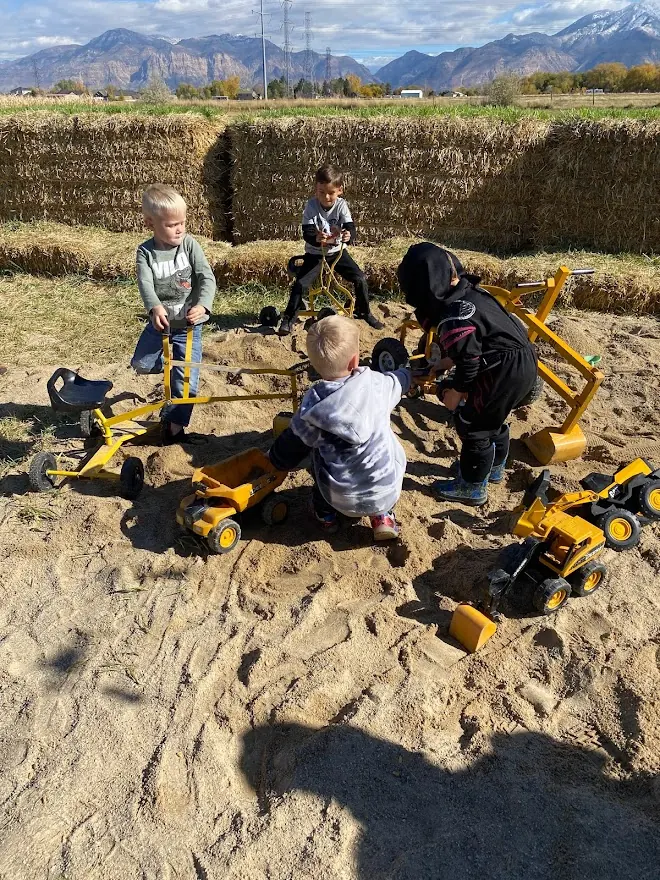 Children working toy diggers and dump trucks in a sandbox ringed by hay bales, with mountains in the distance
