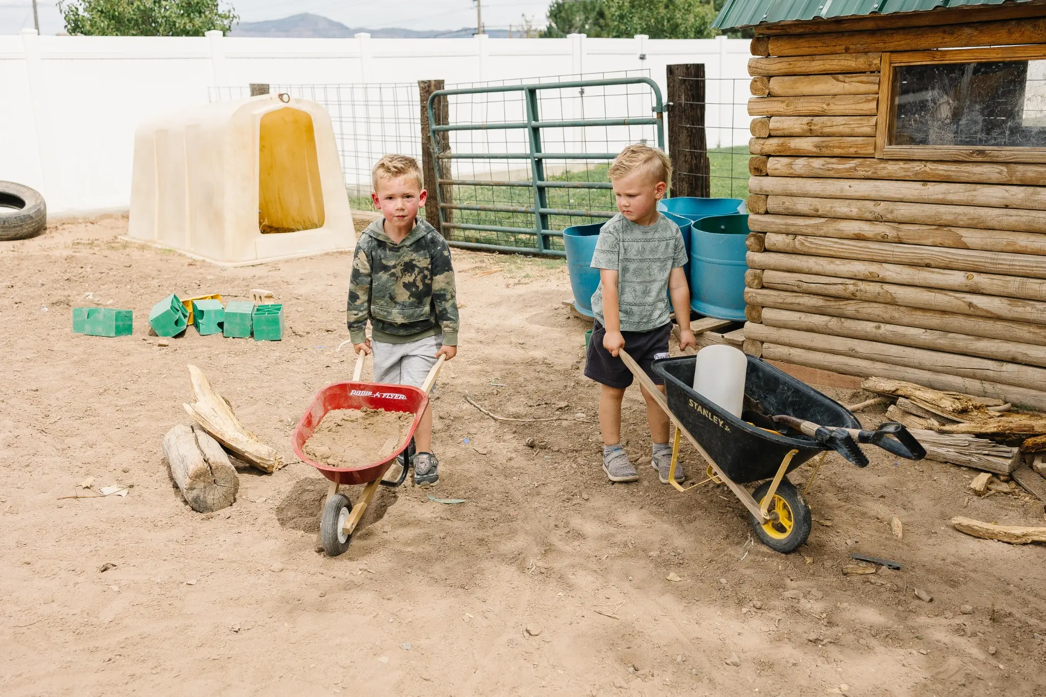 Two young children pushing child-sized wheelbarrows loaded with dirt past the log playhouse in the outdoor classroom