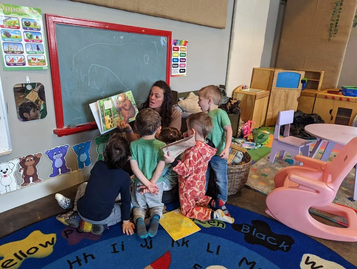 Christi Warr holding up a picture book while a small group of children gather close around her on the classroom rug