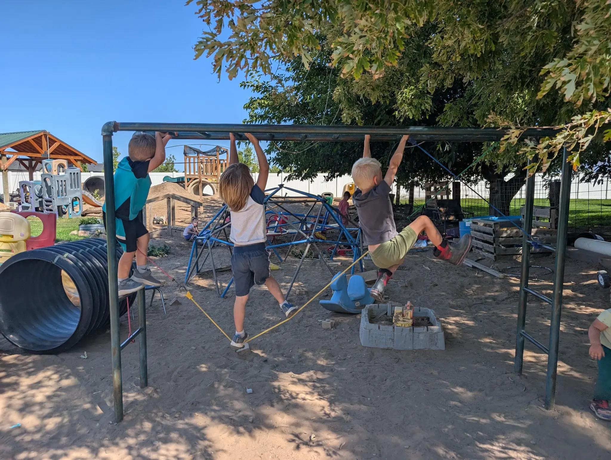 Three children hanging side by side from a tall overhead bar in the sunny outdoor play yard, with a climbing dome, crawl tunnel, and sandbox behind them