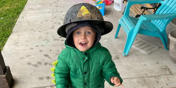 A beaming toddler in a green puffer coat wearing an oversized toy firefighter helmet during pretend play on a covered patio