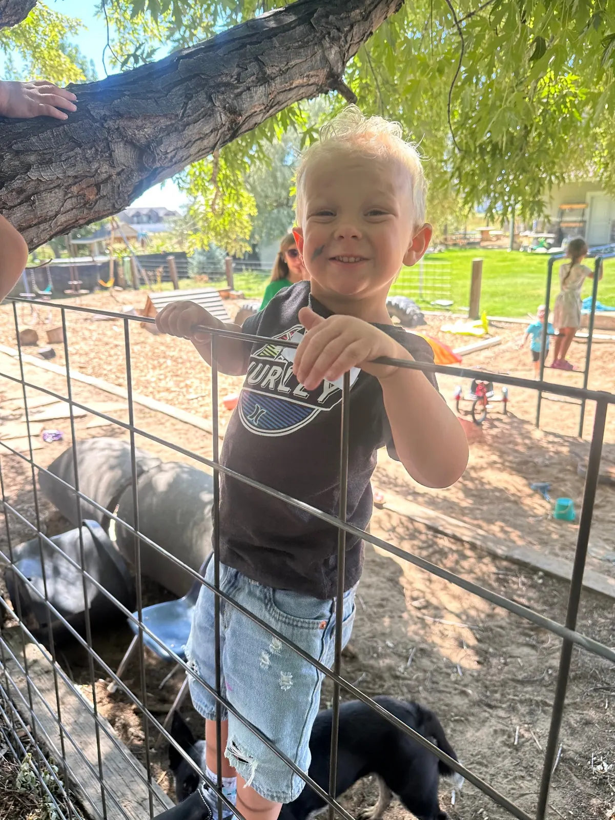 A young child grinning while holding the top rail of a wire fence in the shaded outdoor play yard