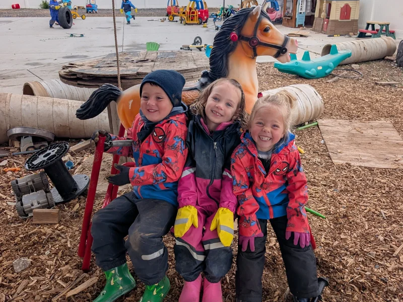 Three smiling children in colorful rain suits sitting together on a toy rocking horse in the outdoor play yard