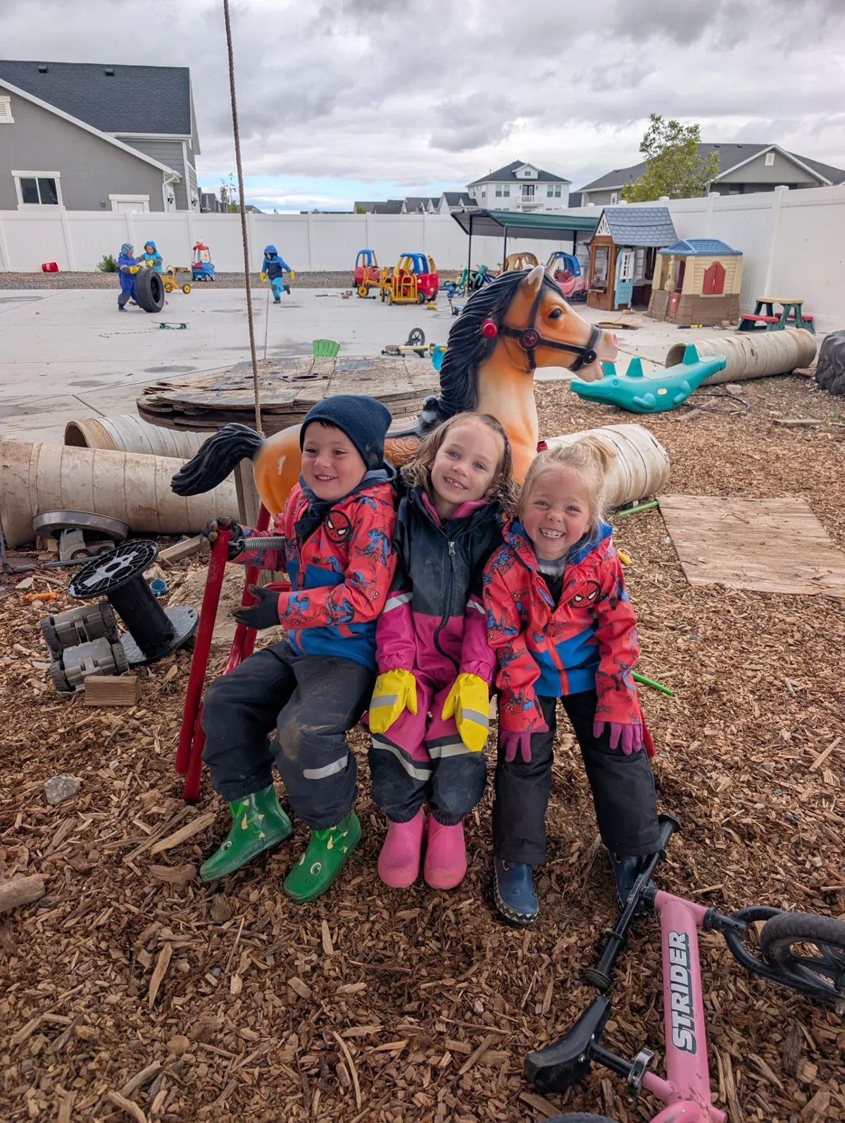 Three bundled-up children squeezed together on a swing in the wood-chip play yard, with a toy horse and ride-on toys behind them