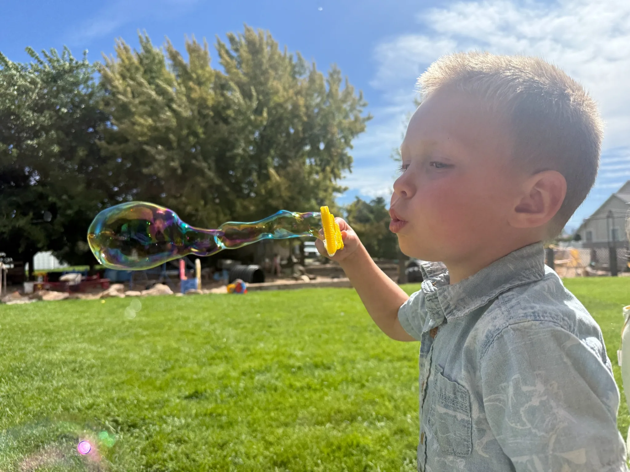 A young child on the sunny lawn blowing a long, shimmering bubble through a yellow wand, with the play yard behind them