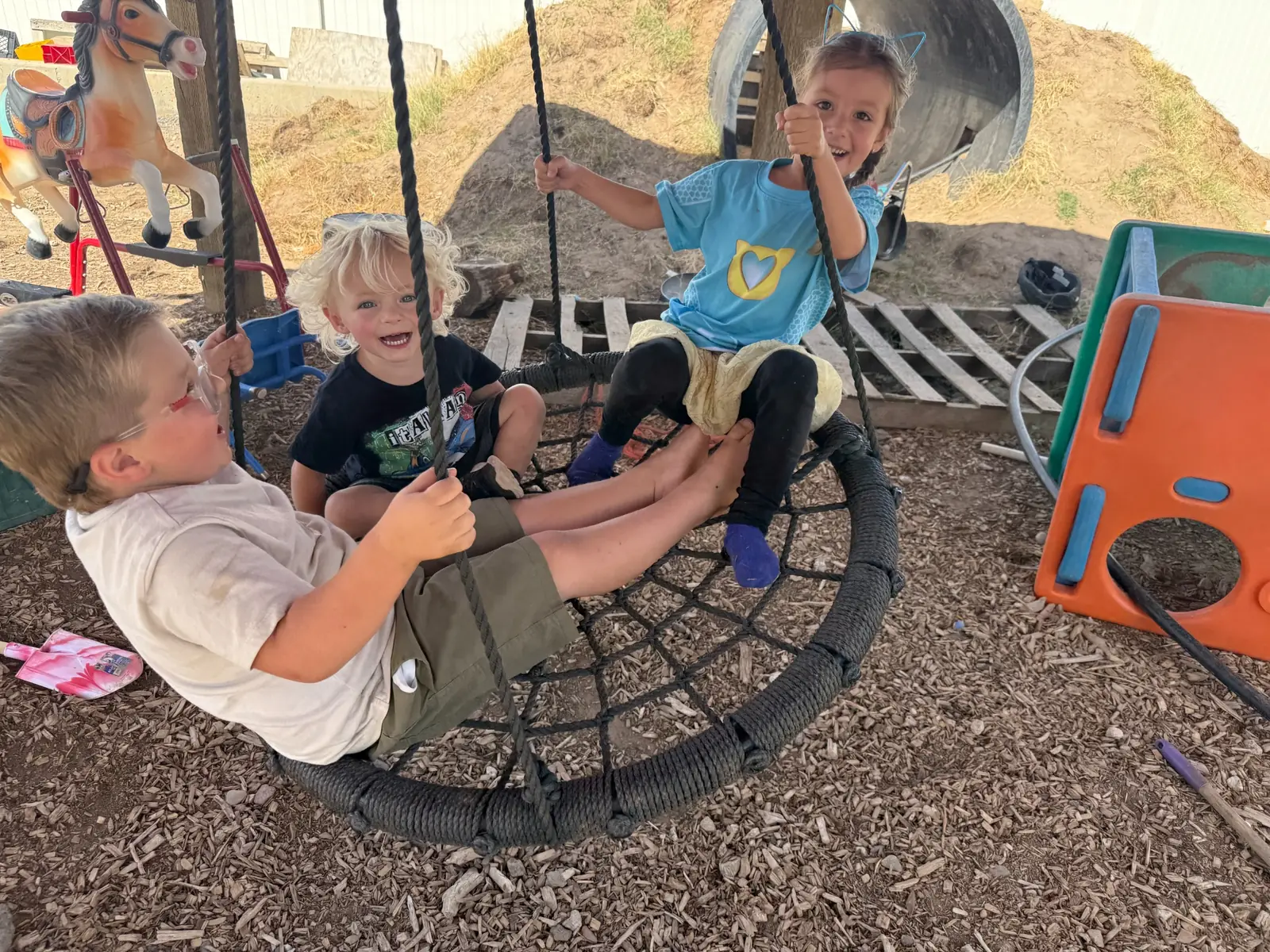 Three laughing children sharing a round rope nest swing, holding the ropes as they sway over a wood-chip play yard