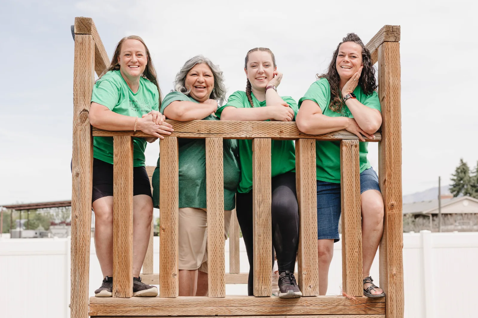 The four Dirt Learning play-based facilitators in green Dirt Learning t-shirts, smiling together at the top of the wooden play structure