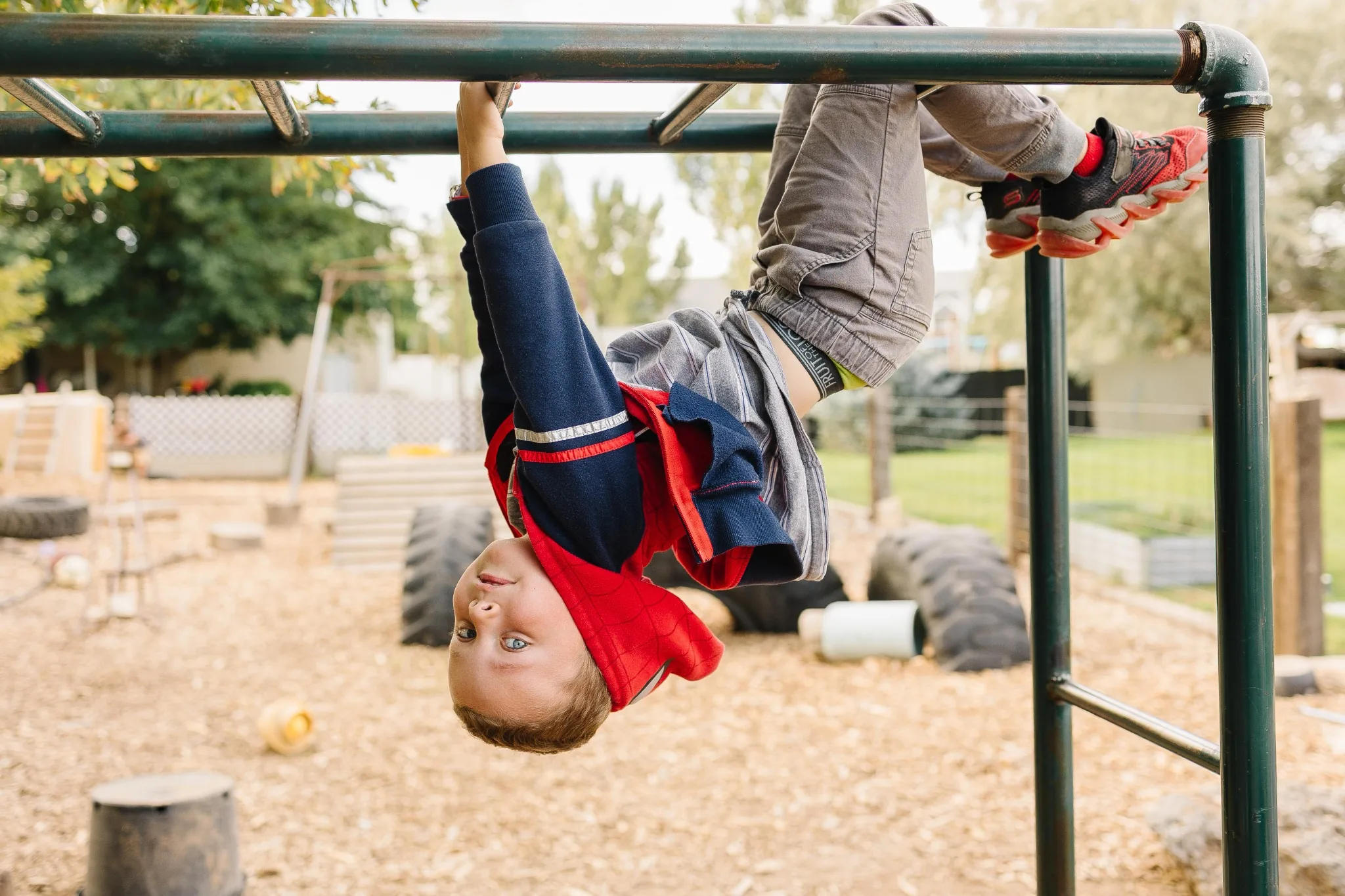 A child hanging upside down by the knees from tall green monkey bars, grinning at the camera above the wood-chip play yard