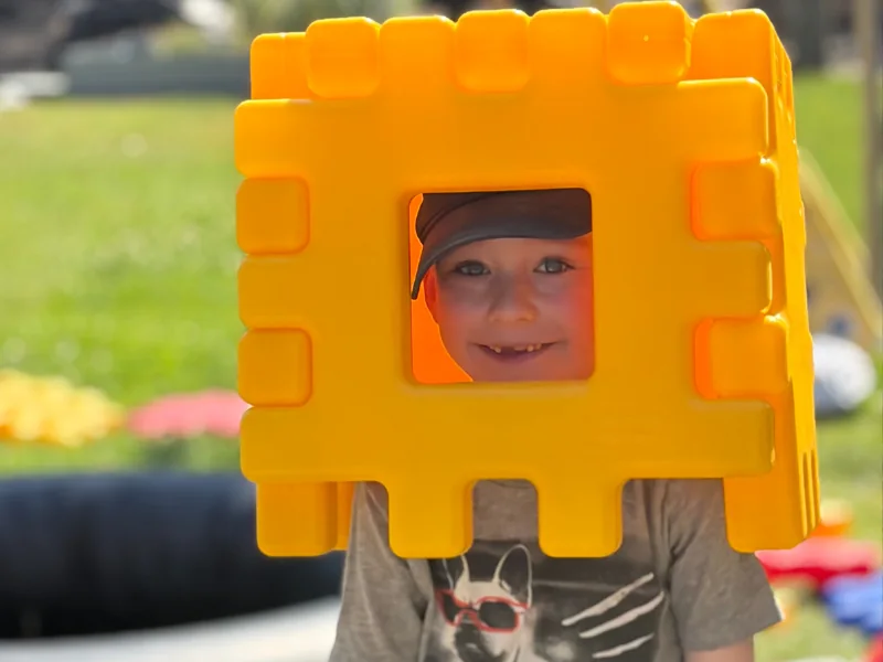 A smiling child peeking through the opening of a large yellow construction block during outdoor play