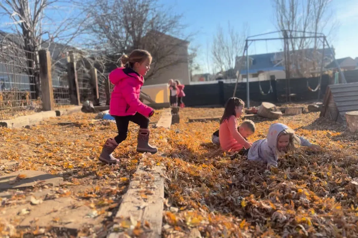 Children romping through a deep drift of golden fall leaves in the sunny outdoor play yard, with swings and bare trees behind them