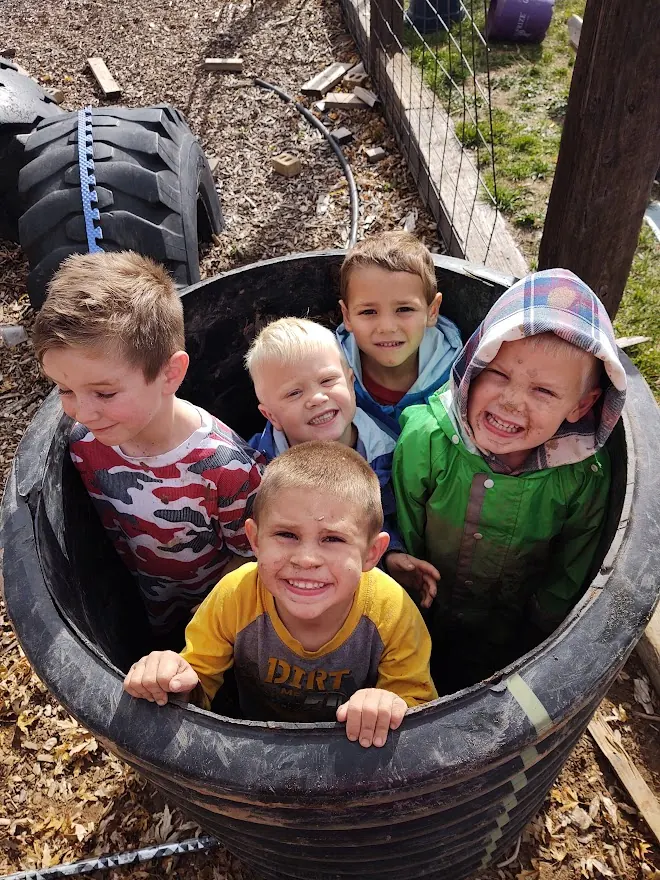 Five smiling children squeezed together inside a big black tube on the wood-chip play yard